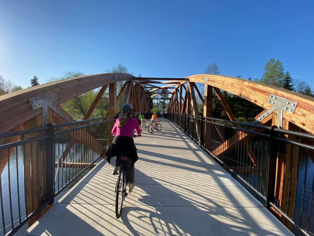 A person in a pink jacket rides a bike on a wooden truss bridge with shadows cast on the pathway.