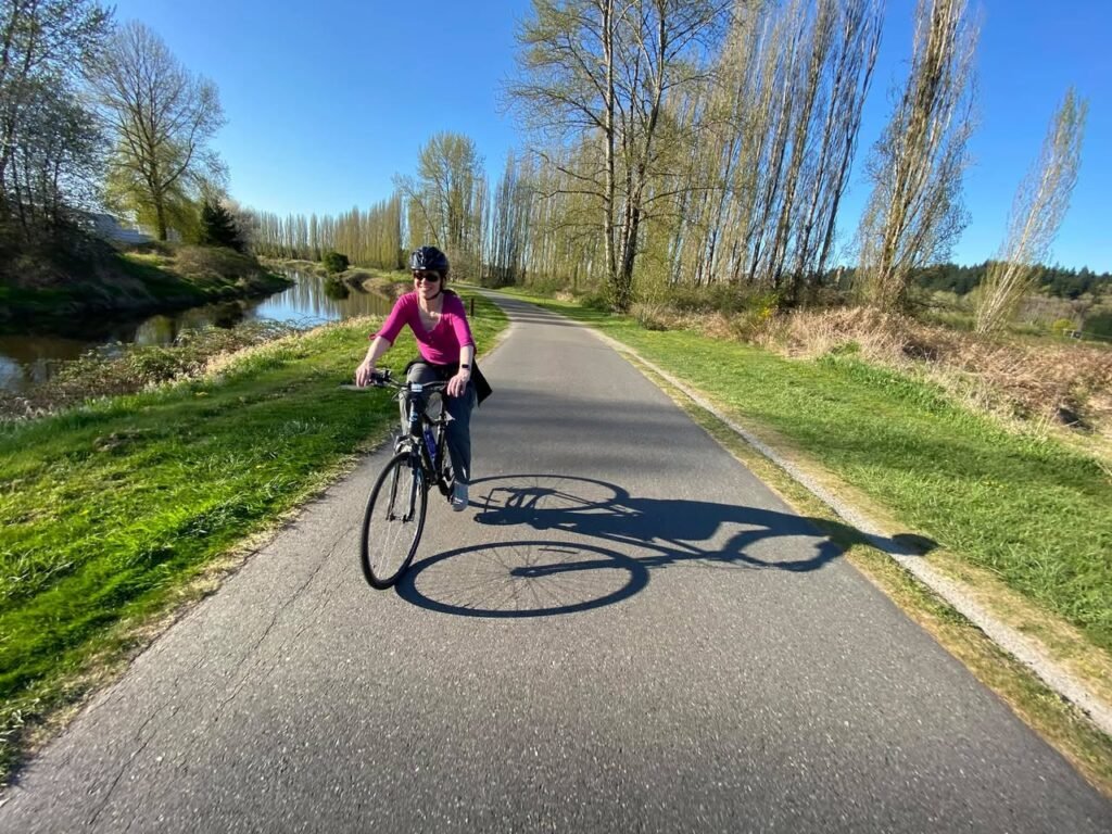 A cyclist rides along a paved path beside a calm river under a clear blue sky, with lush greenery and tall trees lining the route.