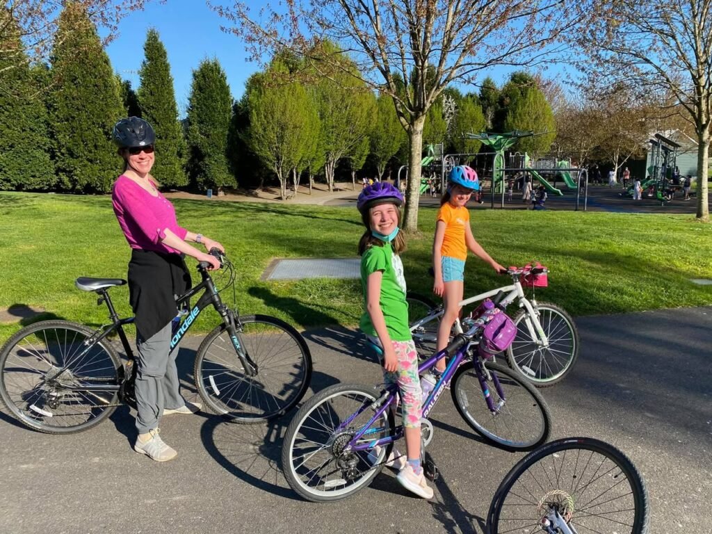 A woman and two children on bicycles in a sunny park with tall evergreen trees. A close-up view of one individual wearing a pink top and cycling gear.