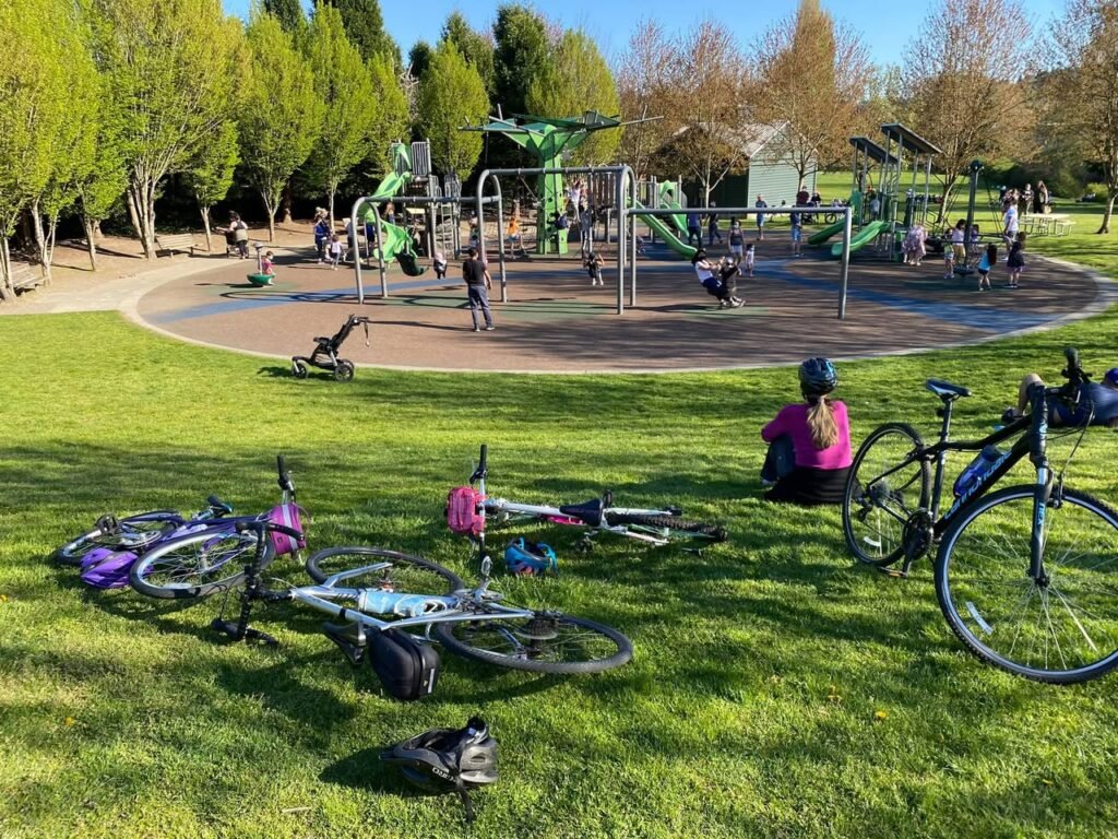 A bright, sunny day at a lively park playground with cyclists enjoying the green space and bike path.