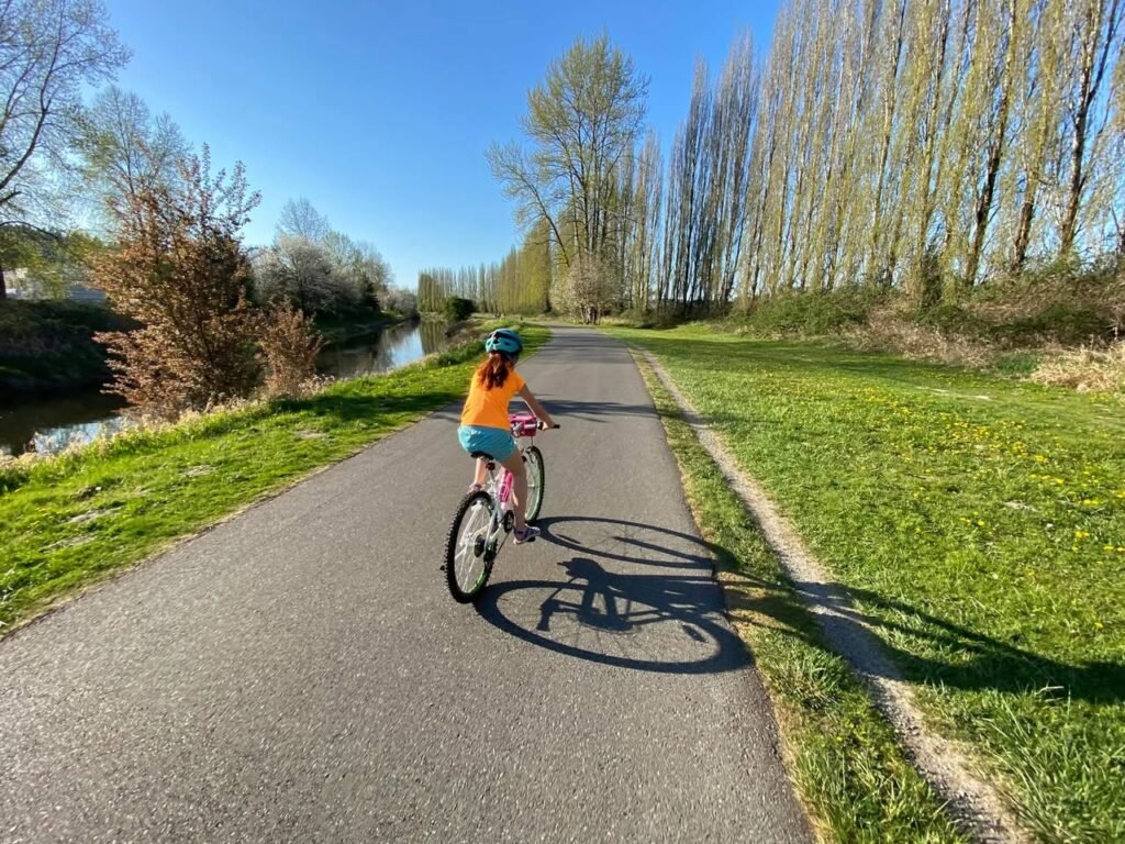 A person wearing an orange shirt and helmet rides a bicycle along a scenic paved path lined with budding trees and a canal under a clear blue sky.