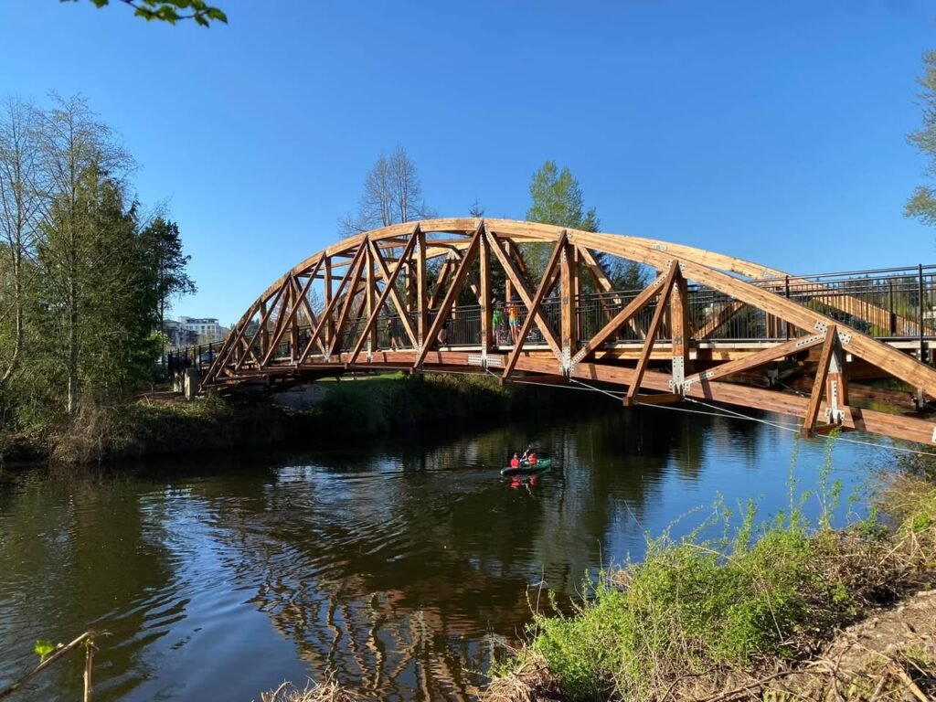 A rust-colored truss bridge spans a calm river under a clear blue sky, with surrounding trees and greenery adding to the scenic beauty.