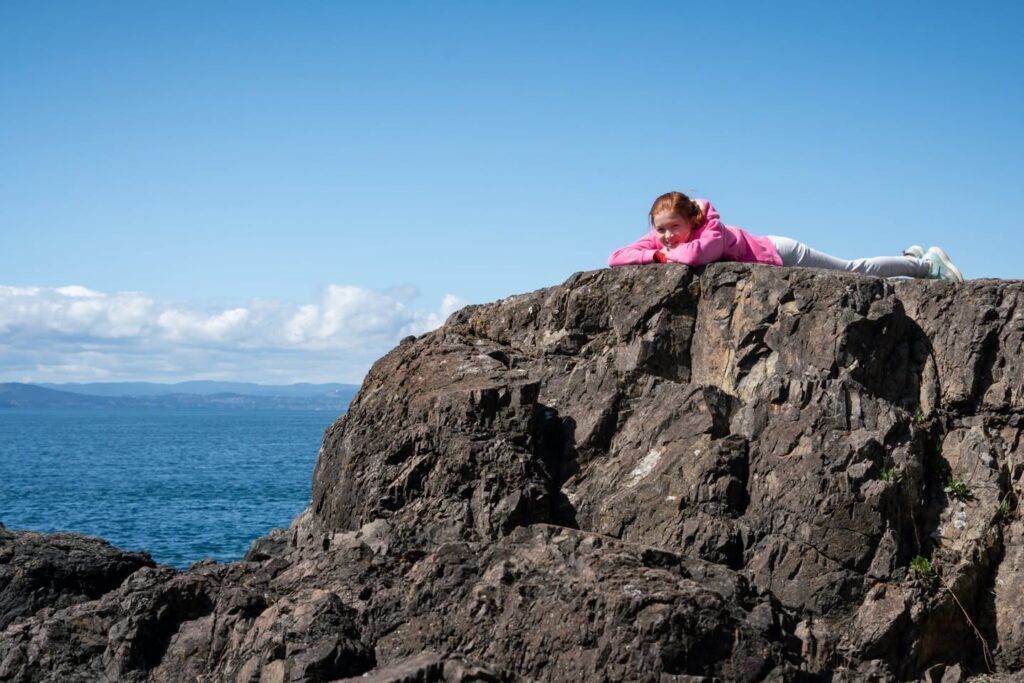 A person in pink clothing reclines on rugged coastal cliffs overlooking the vast, blue ocean under a clear sky.