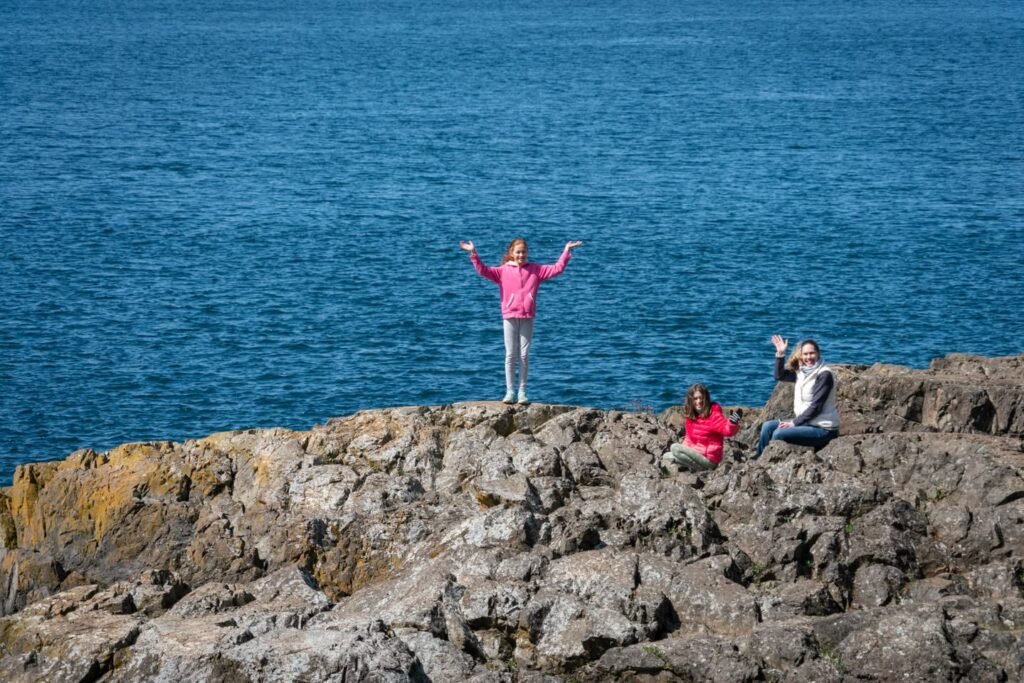 A person standing on a rocky outcrop with arms raised against a backdrop of deep blue ocean water.
