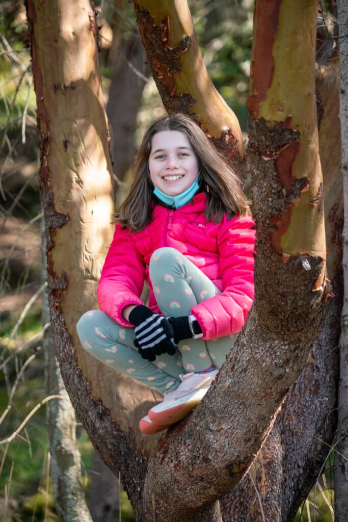 A person sitting nestled in the fork of a madrone tree wearing a bright pink puffer jacket.