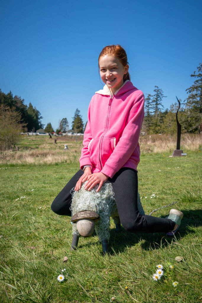 A smiling person in a pink hoodie kneels on grass in a sunny park, petting a gray dog wearing a scarf.