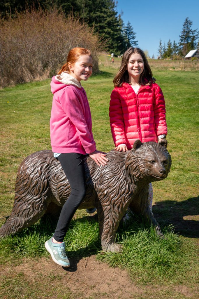 Two people posing next to a realistic looking bear sculpture made of twigs in a grassy field.