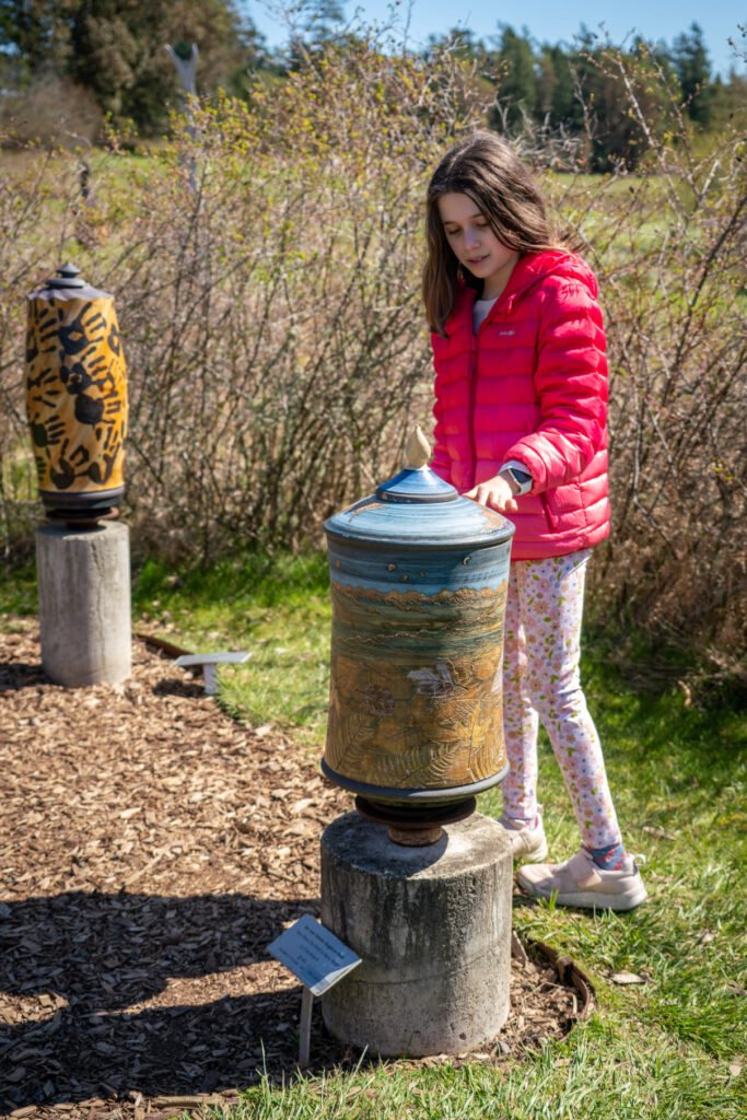 A person in a red jacket examines a ceramic sculpture at an outdoor gallery. Natural surroundings of grass and bushes are visible in the background.