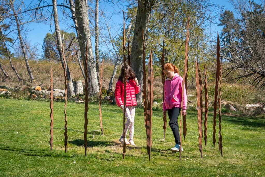 A whimsical outdoor art installation made of twisted willow branches. Visitors walk through and around the natural sculpture on a sunny day.