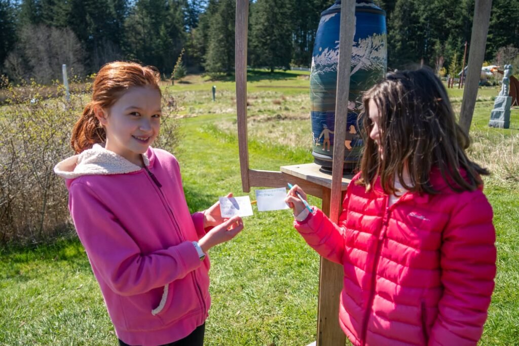 Two children in pink jackets hold cards at a rustic outdoor kiosk.
