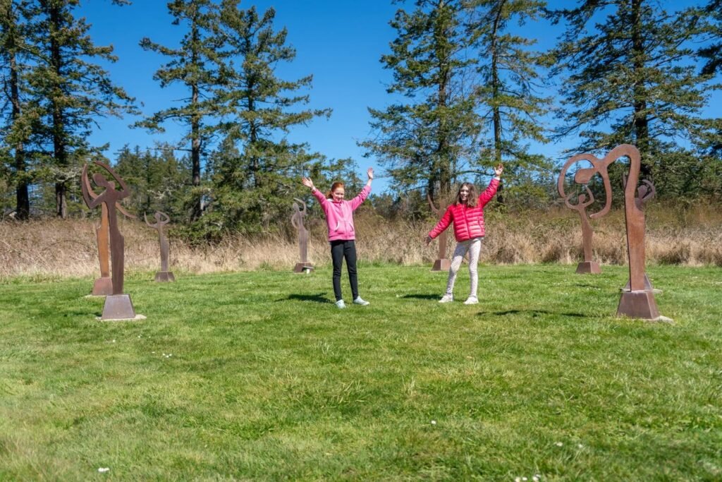 Two people pose with arms raised in a grassy field surrounded by tall pine trees and abstract rust-colored metal sculptures under a clear blue sky.