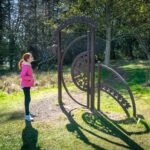 A metal spiral sculpture on a forest floor with a child standing nearby on a sunny day.