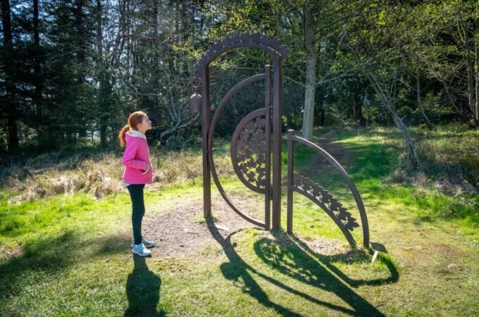 A metal spiral sculpture on a forest floor with a child standing nearby on a sunny day.