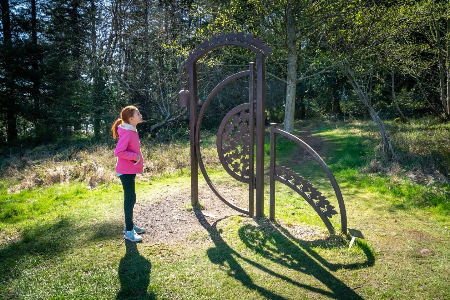 A metal spiral sculpture on a forest floor with a child standing nearby on a sunny day.