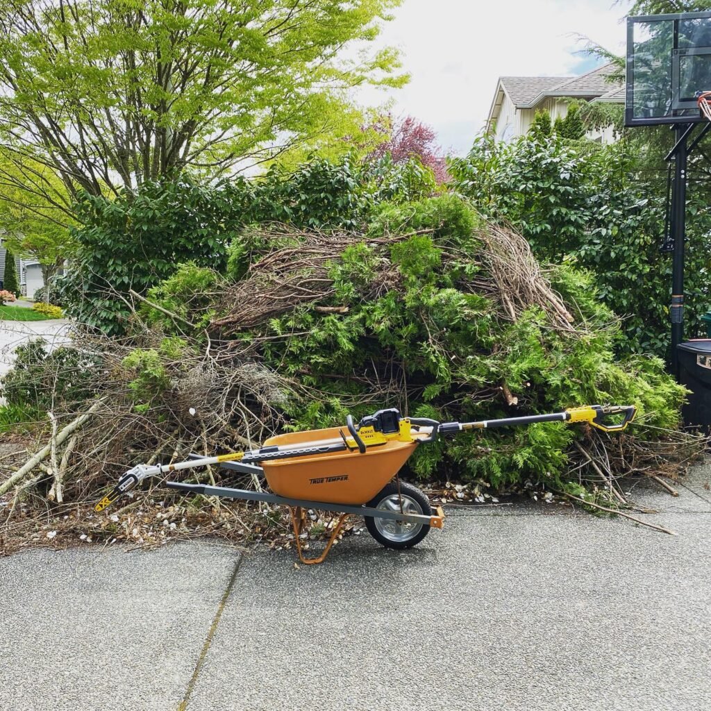 An orange wheelbarrow-style yard waste wagon with a shredder attachment sits in front of a large pile of fresh green branches.