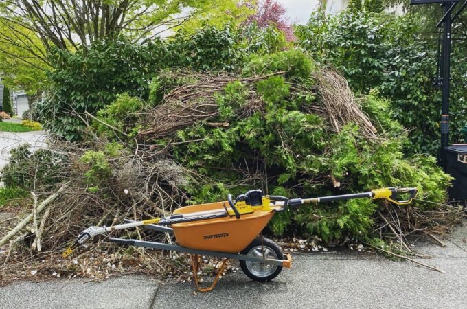 An orange wheelbarrow-style yard waste wagon with a shredder attachment sits in front of a large pile of fresh green branches.