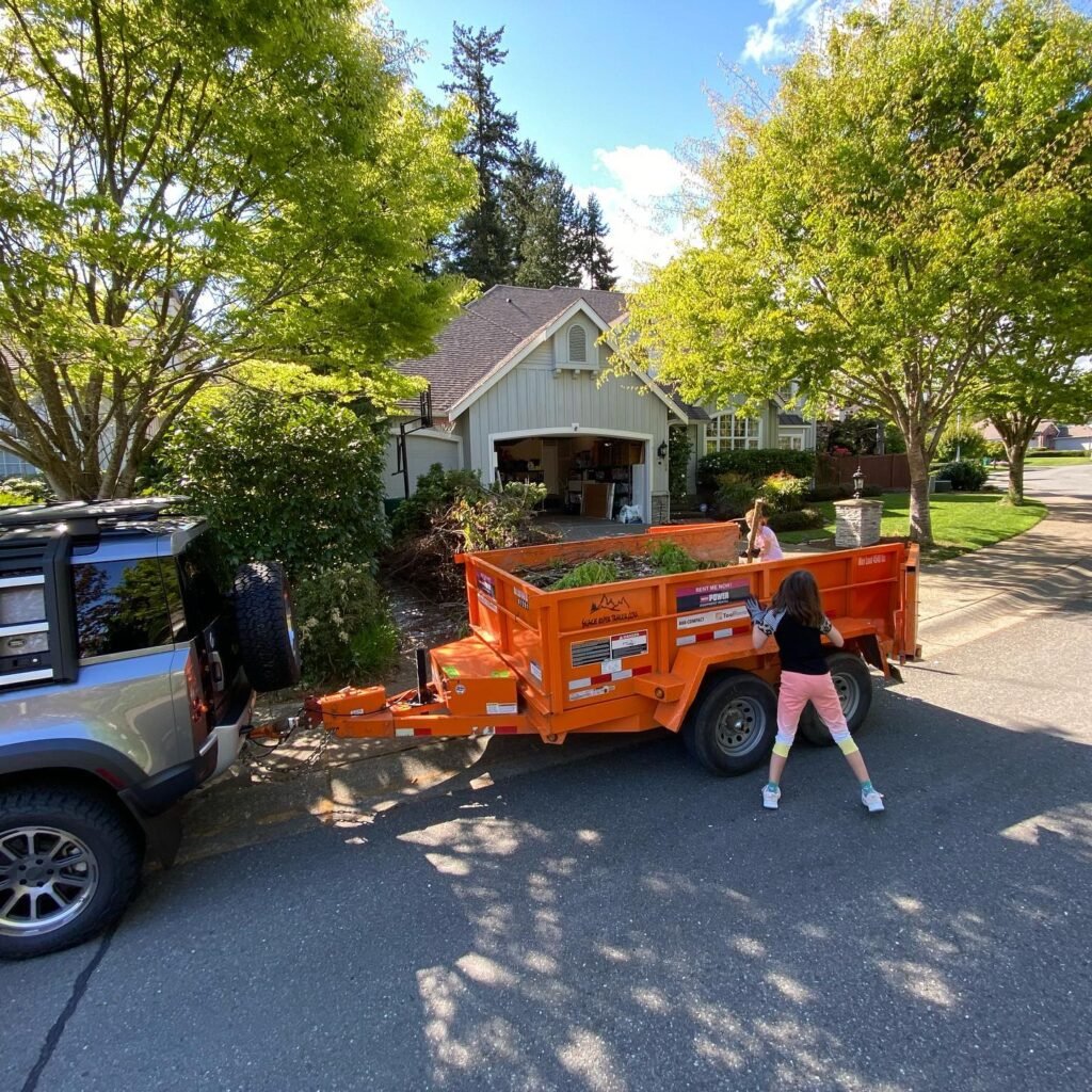 A bright orange trailer filled with landscaping material is pulled by a vehicle in front of a house in the Pacific Northwest.