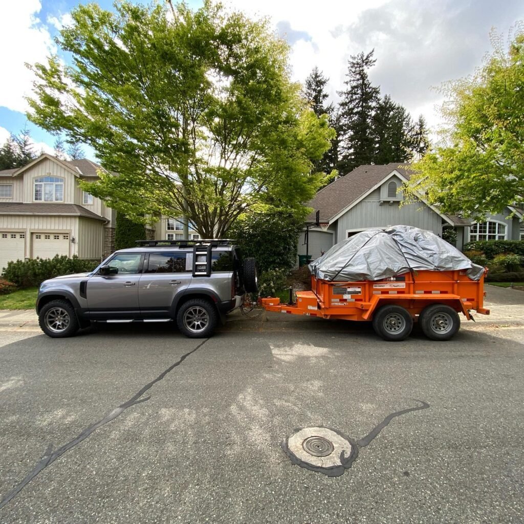 A silver SUV is shown towing an orange trailer with a covered jet ski on a residential street. Lush trees and a house are visible in the background.