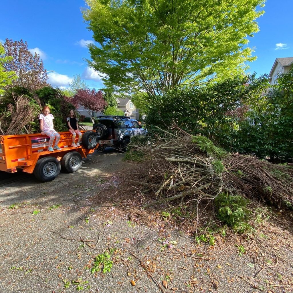A utility trailer loaded with yard waste sits in a driveway surrounded by green trees under a blue sky. A large pile of branches rests nearby on the gravel.