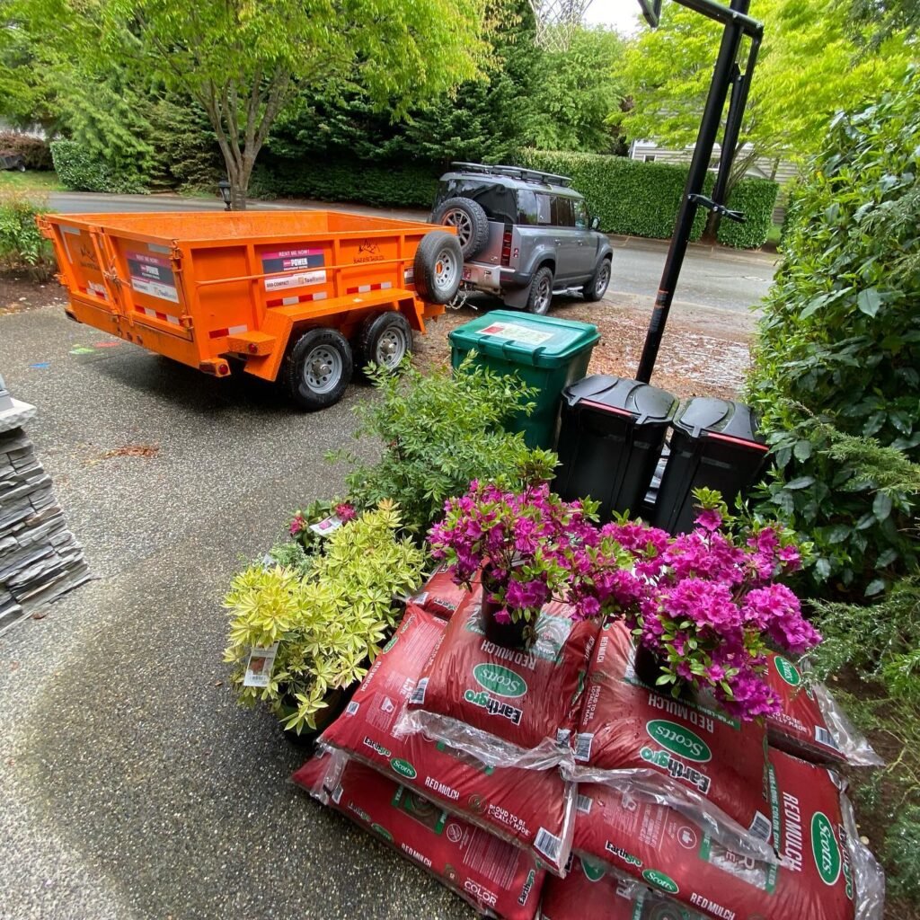 An orange dump trailer sits in a driveway near bags of soil and fresh spring plantings. Vivid pink blooms and yellow-green shrubs are arranged atop red sacks in the Pacific Northwest setting.