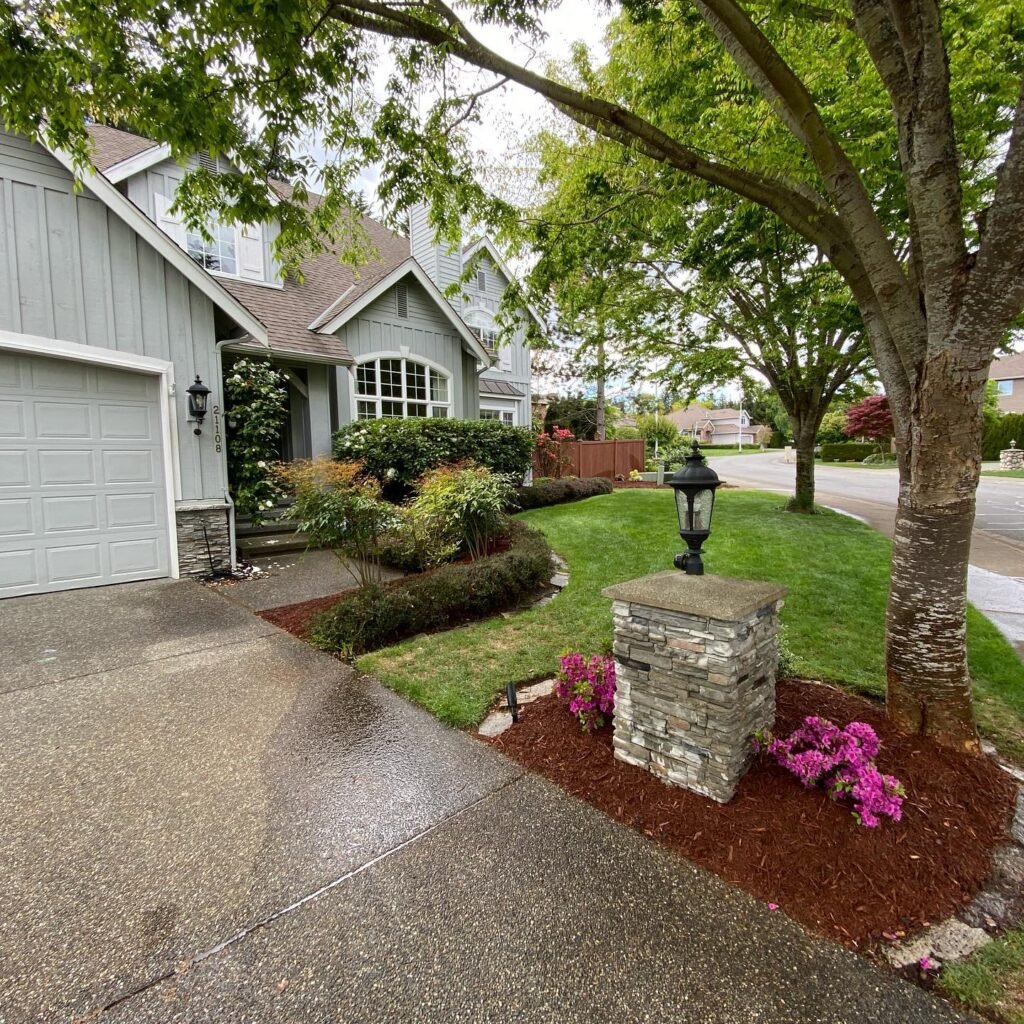 A view of a gray house's exterior with a newly stripped concrete driveway, surrounded by trees, shrubs, and purple flowers.