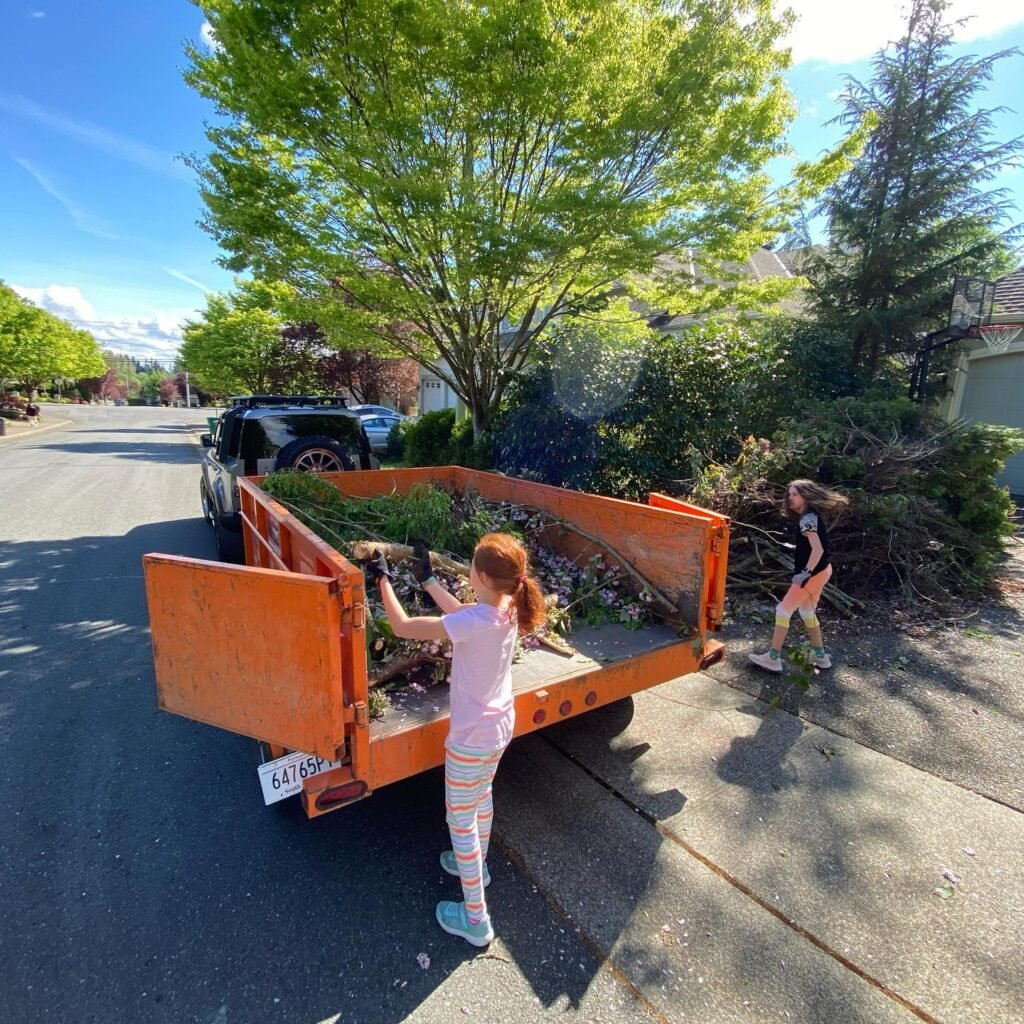Children assist in cleaning yard debris on a sunny day in the Pacific Northwest. A pickup truck's tailgate is open on a quiet suburban street.