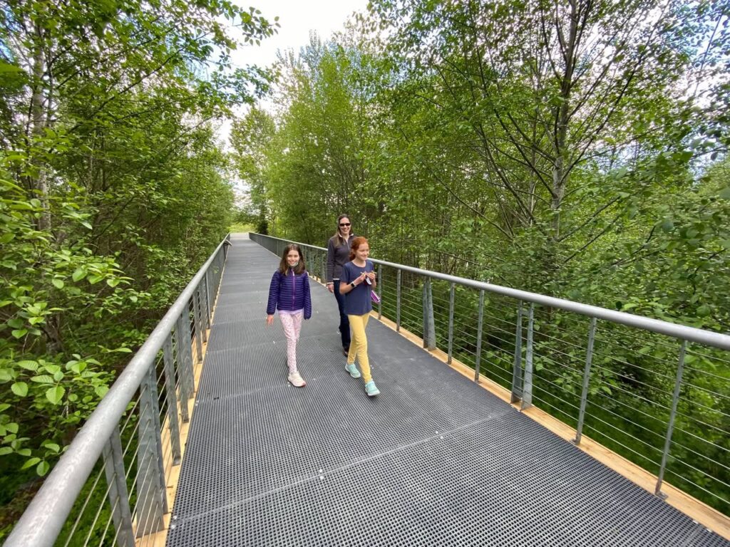 A family of three strolls along a metal canopy bridge surrounded by lush green trees.