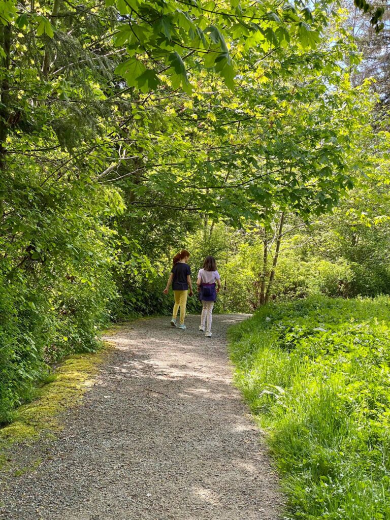 Two people walk along a sun-dappled gravel path surrounded by dense green foliage in a park.