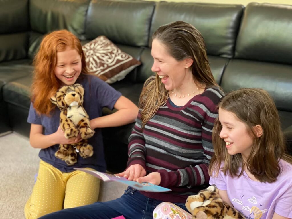 A woman and two young girls, each holding a stuffed cheetah toy, exploring craft items on the floor. The image captures a lively and engaging family moment.