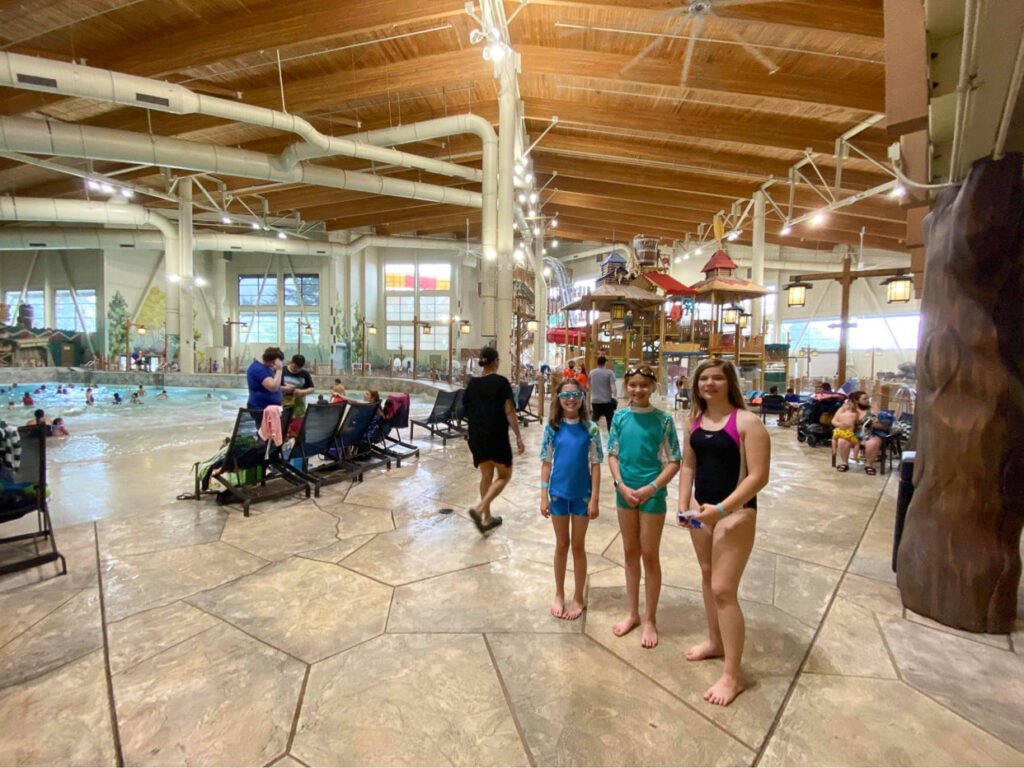 Children in swimsuits pose by the pool at a spacious indoor water park. The high wooden ceiling features exposed beams and a large tree-like column to the right.