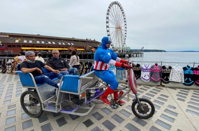 A person dressed as Captain America rides a custom tricycle with two passengers near a waterfront pier. A large Ferris wheel stands in the background under an overcast sky.
