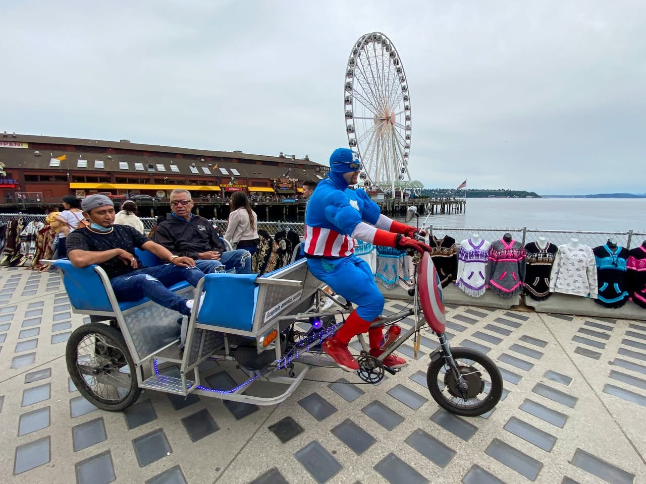 A person dressed as Captain America rides a custom tricycle with two passengers near a waterfront pier. A large Ferris wheel stands in the background under an overcast sky.