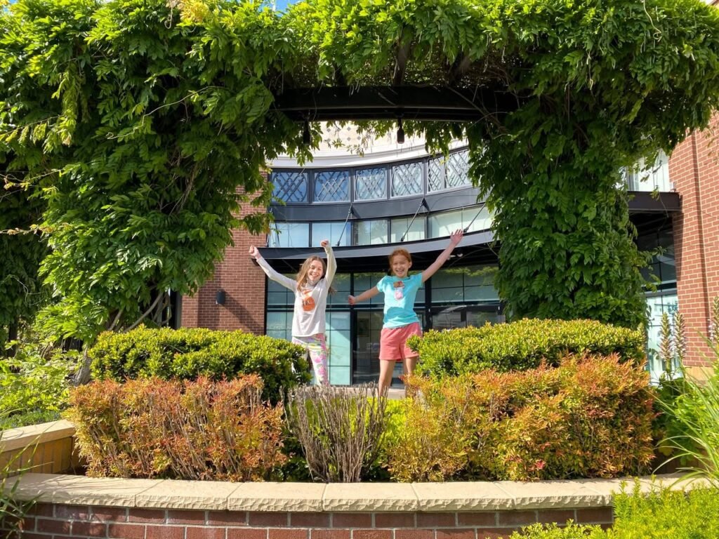 Two young children raise their arms in excitement on a brick ledge in a beautifully landscaped garden. A leafy ivy-covered archway frames a modern brick building in the background.