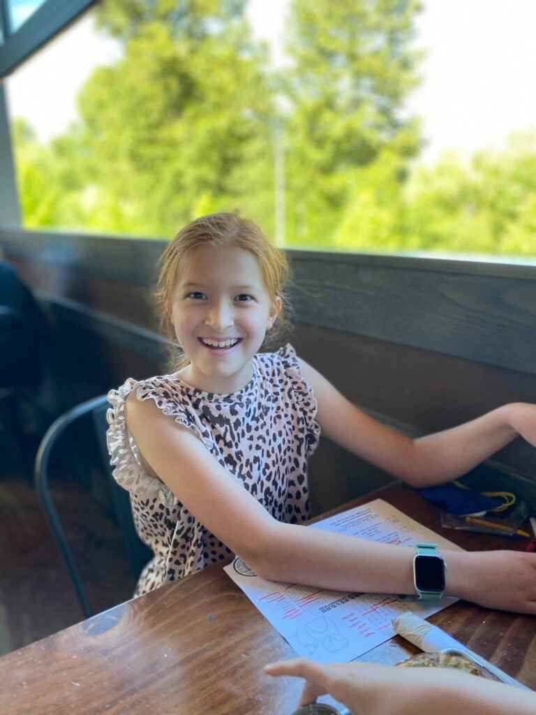 A smiling child sits at a wooden table outdoors with bright green trees in the background. The person is wearing a patterned sleeveless top and a smartwatch.
