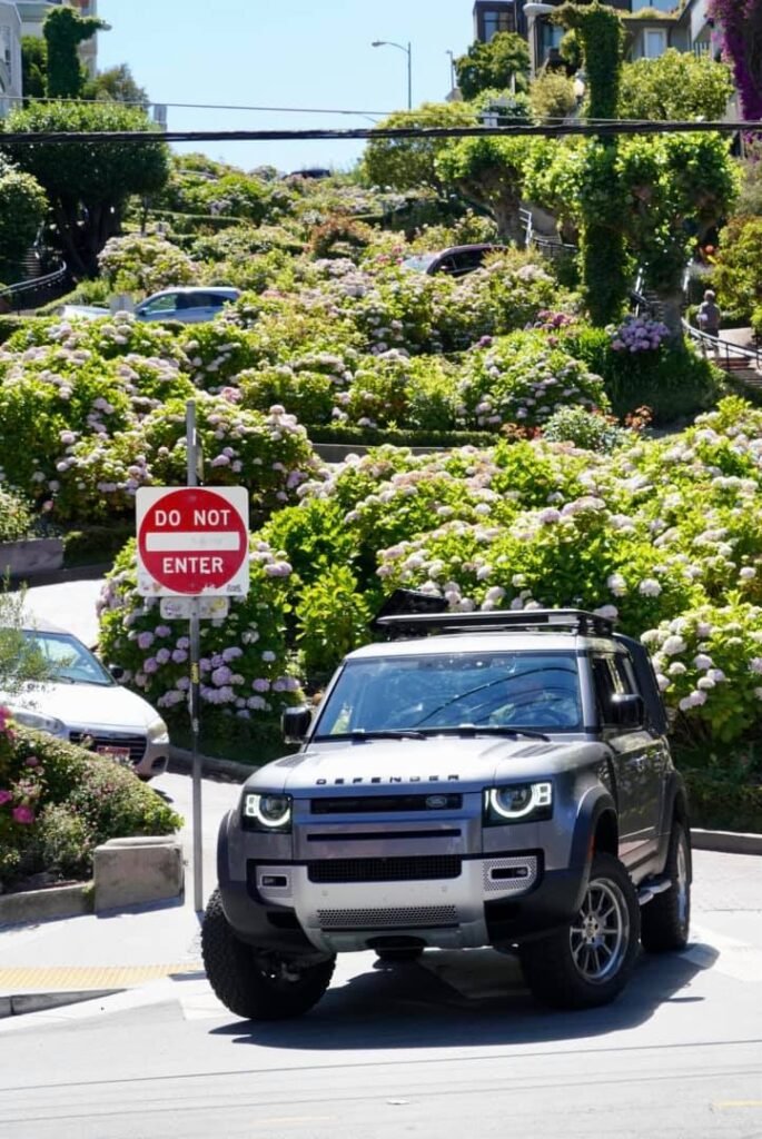 An SUV drives past a do not enter sign on a vibrant, flower-filled hillside street during travel.