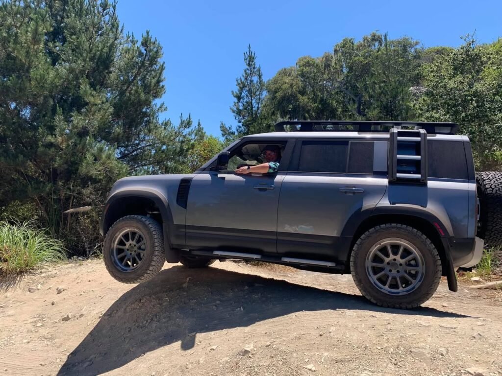 A grey SUV is navigating a dirt trail amidst lush green trees, with a close-up of its tire and roof rack shown against a clear blue sky.