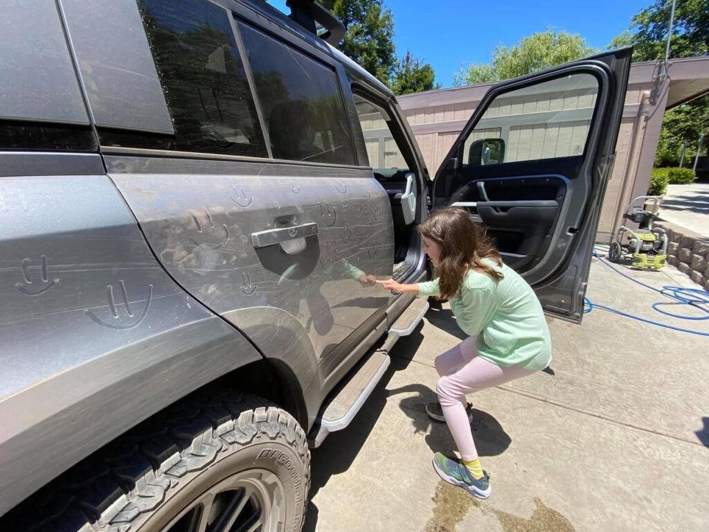 A child in pink pants and a green shirt crouches by a silver SUV, wiping the lower side panel. A pressure washer sits in the background near a garage on a sunny day.