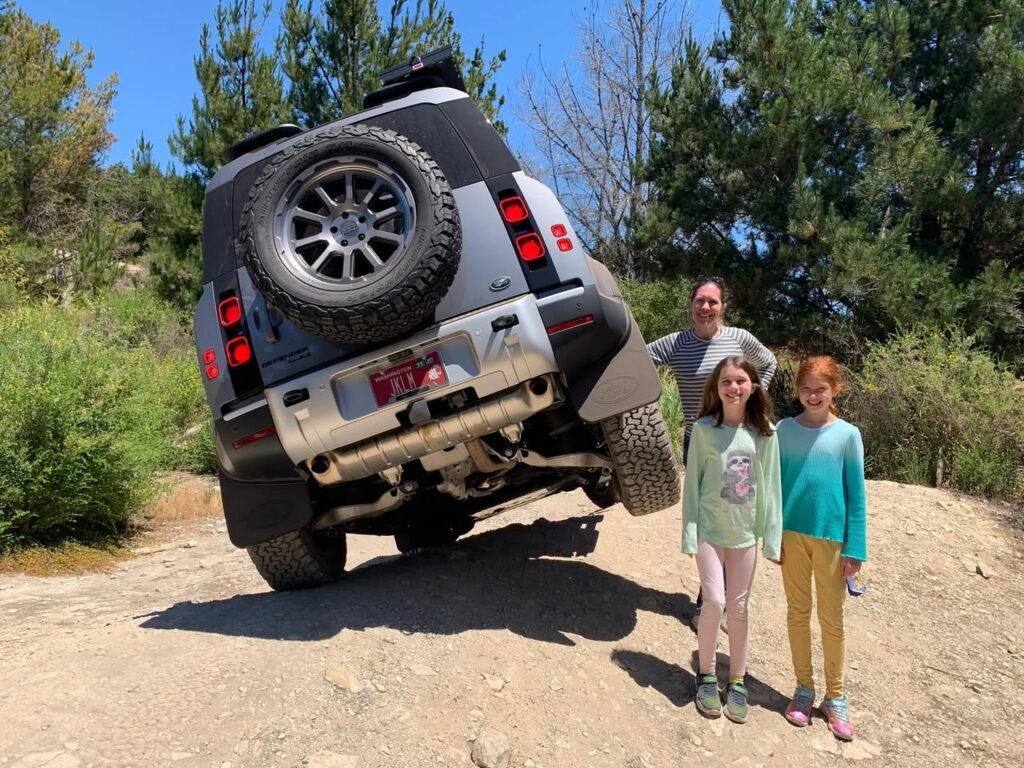 A silver Jeep Wrangler is tilted forward on a dirt hilltop with a man and two people walking beside it in a sunny outdoor setting. The image features multiple angles of the vehicle and the people.