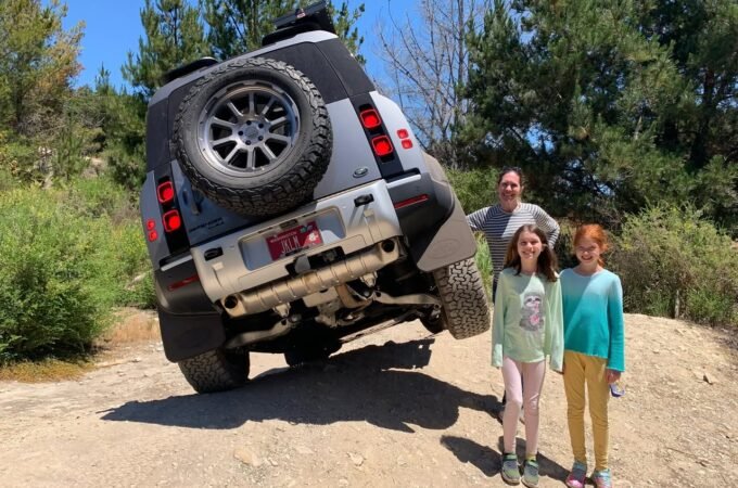 A silver Jeep Wrangler is tilted forward on a dirt hilltop with a man and two people walking beside it in a sunny outdoor setting. The image features multiple angles of the vehicle and the people.