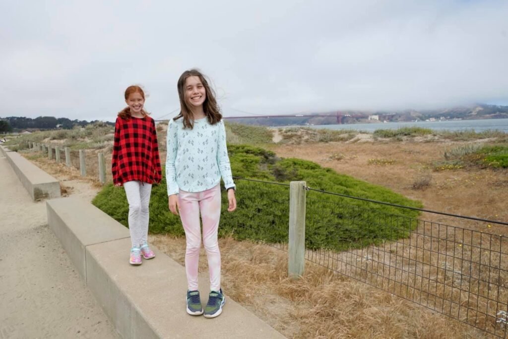 A family stands on a boardwalk overlooking a coastal landscape with dry grass and a distant view.