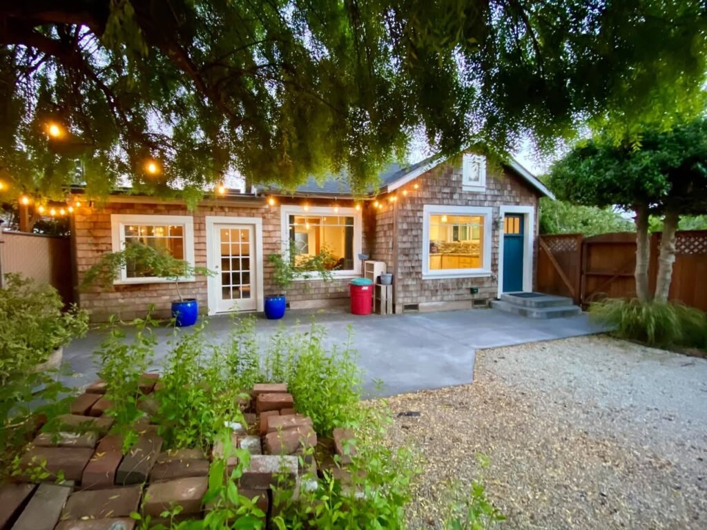 A cozy brick backyard with a concrete patio, garden beds, and string lights. Colorful buckets and lush greenery enhance the outdoor setting.