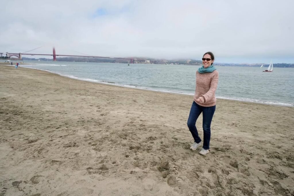 Person standing on sandy beach with Golden Gate Bridge and boats in the background on a cloudy day.