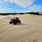 Bandon Beach An off-road vehicle rides over large sand dunes with a line of trees in the background. The sky is bright blue with wispy clouds.