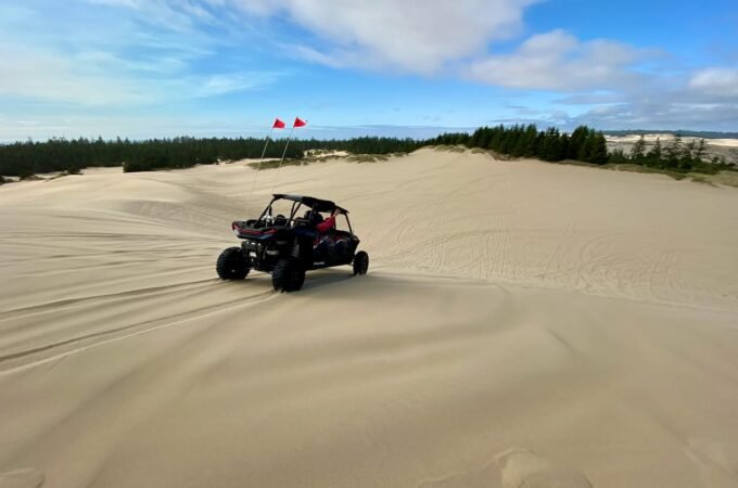 An off-road vehicle rides over large sand dunes with a line of trees in the background. The sky is bright blue with wispy clouds.