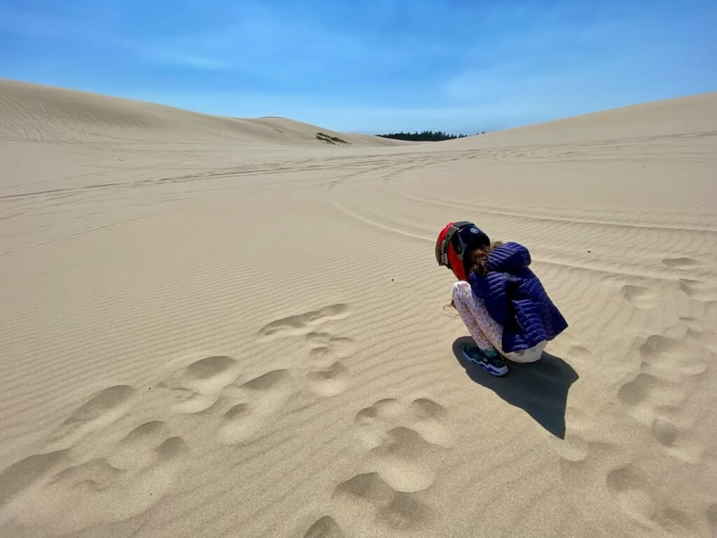A person wearing a helmet crouches on Sandoval Beach sand dunes while footprints trail across the rippled, golden sand under a clear blue sky.