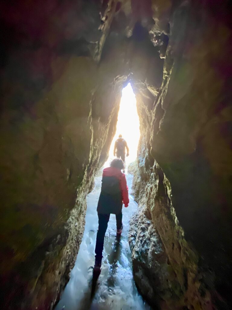 Hikers walking on a puddled canyon path with bright sunlight streaming from the opening.