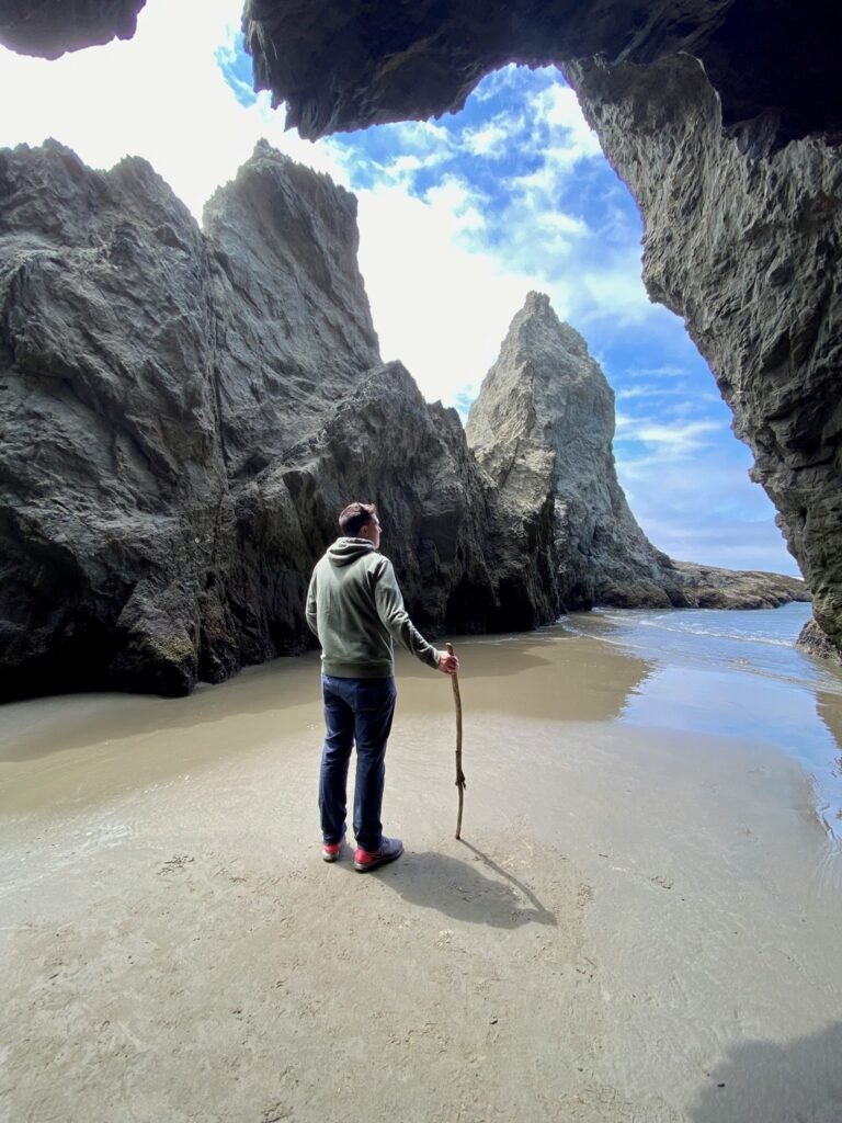 A person stands on wet sand below a large rock arch looking out at tall sea stacks and the ocean under a blue sky.