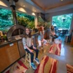 A cozy indoor dining space at Barefoot Ranch in Portland with large windows opening to lush greenery. People sit on stools and look out at the nature view.