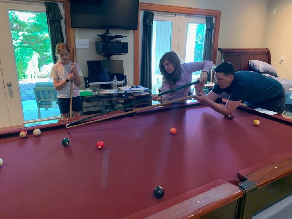 Friends and family playing pool on a red table in a home living room. A casual indoor gathering featuring billiards and a bedroom in the background.