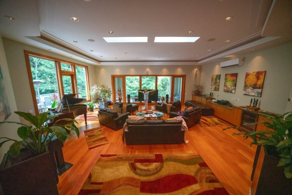 A spacious living room featuring wooden floors, a patterned rug, and large windows overlooking greenery.